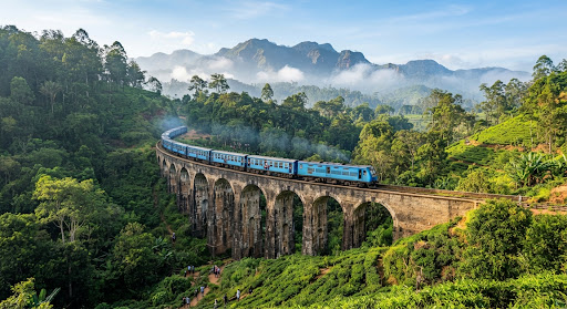 The world-famous Nine Arch Bridge in Ella, Sri Lanka with the iconic blue scenic train crossing over stone arches surrounded by dense emerald jungle and tea fields, morning tropical mist rising from mountain ridges, editorial warm lighting.