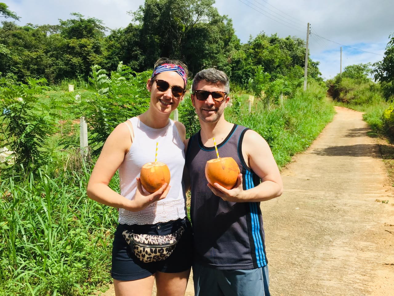 A smiling couple holds fresh king coconuts with straws along a rural dirt road.
