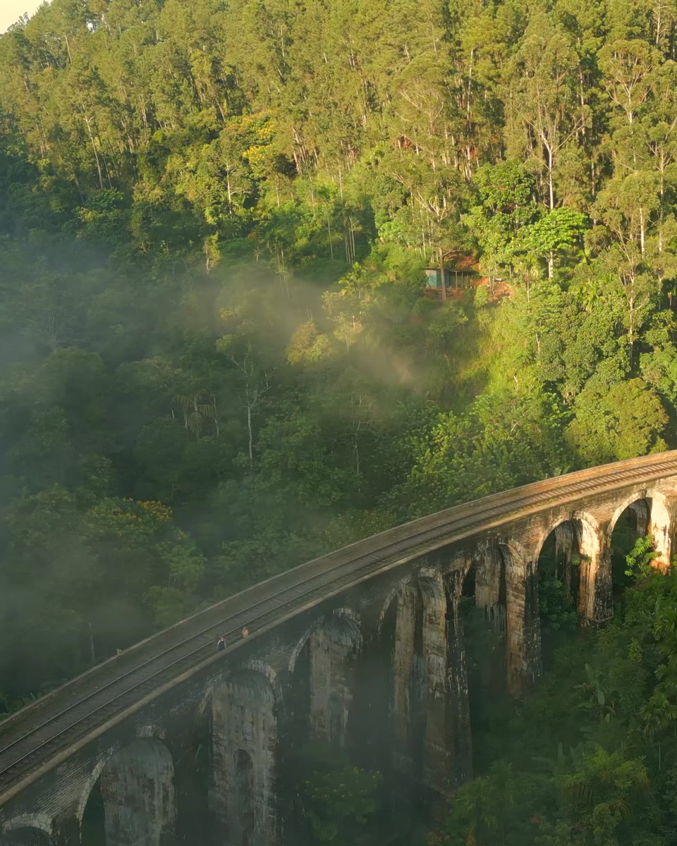The stone arches of Ella's Nine Arch Bridge curving through a jungle-covered valley as morning mist drifts through the sunlit trees.