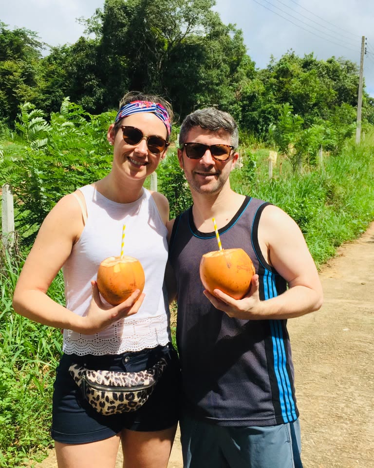 Two smiling travellers holding fresh king coconuts with straws at a leafy roadside stop in Sri Lanka.
