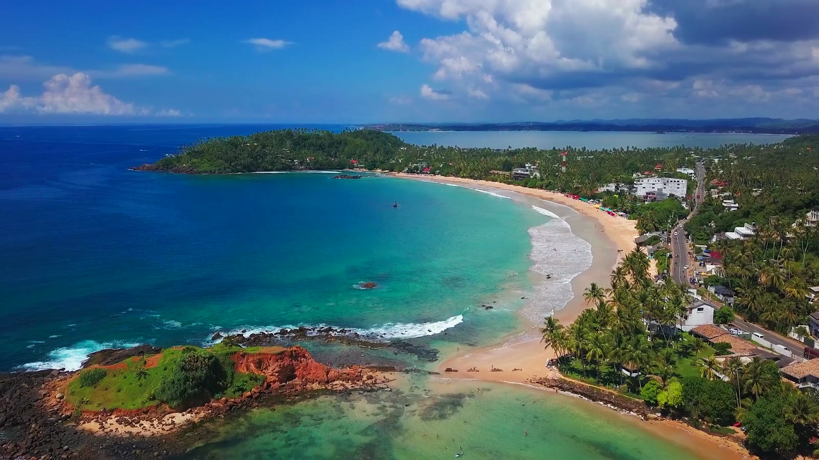Aerial view of Mirissa bay on a sunny day: a curved golden beach, turquoise water, palm-lined shore and a red-rock headland under a bright blue sky.